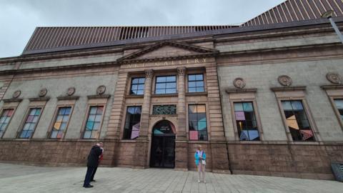 An exterior view of the front of Aberdeen Art Gallery. A stone building with columns and carvings, under a partly cloudy blue sky. The words “Art Gallery” are engraved in gold letters above the main entrance. A man and a woman are standing outside the building.