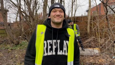 Man in a black hat, black woollen hoodie and bright yellow hi-vis smiling.