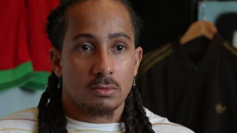 A head-and-shoulders image of Tyler Attwood who is looking serious. He has long dark brown hair styled in dreadlocks. There are sports tops on hangers behind him.