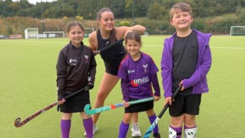 Two small girls and a boy, dressed in black and purple hockey kits, hold hockey sticks up as they smile at the camera. A woman dressed in a black sports vest and shorts, stands behind them and smiles as she holds her hockey stick up high.