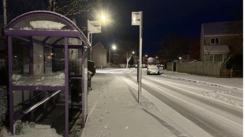 An empty bus stop on the side of a residential road with houses and cars all covered in snow