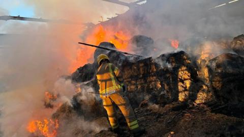 A firefighter pictured from behind in a barn, stood in front of hay which has flames coming out of it and smoke billowing out. The roof of the building has been burnt away.