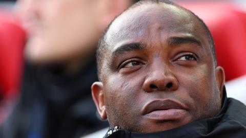 A close-up headshot of Benni McCarthy looking to his left with a high black collar