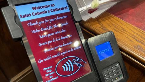 A contactless donation terminal on a wooden surface inside Saint Columb’s Cathedral, showing a welcome screen thanking visitors for donations in multiple languages and instructing users to tap the screen, with a card payment keypad beside it.
