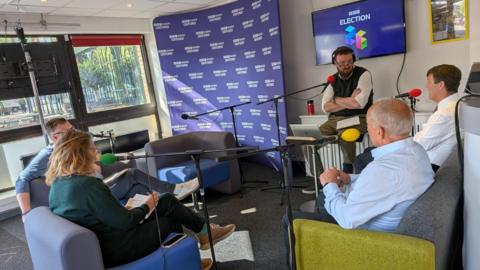 Four people sat on sofas in front of microphones. The presenter is sat on a chair. Behind him is purple BBC election branding.