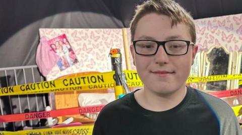 Aston, standing in a drama studio, looking at the camera, wearing glasses, with short hair, he has on a dark T-shirt and is standing in front of a stage set that has caution tape across it. 