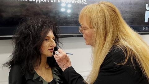 Woman with long blonde hair applies make-up to a woman with long dark hair and a false nose