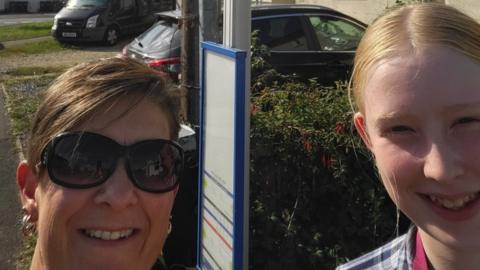 Mel Brown and her daughter, Ruby. They are stood by a bus stop and smiling at the camera.