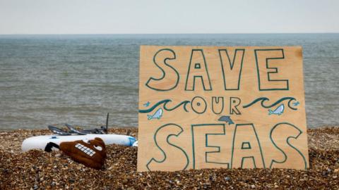 A handwritten sign on the beach at Hythe reads "Save our seas" as part of a protest organised by Surfers Against Sewage on 18 May 2024