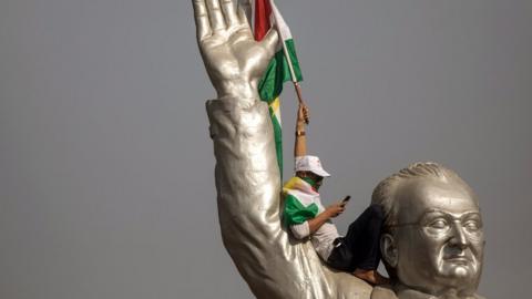 A supporter of Kurdish independence waves a flag while sat on the shoulder of a large stadium statue in Erbil.
