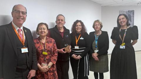 Paul Newman is wearing a black bodywarmer over a claret polo shirt. He is smiling and holding his award. There are three women to his left and a man and a woman to his right. They are all smartly-dressed and smiling.