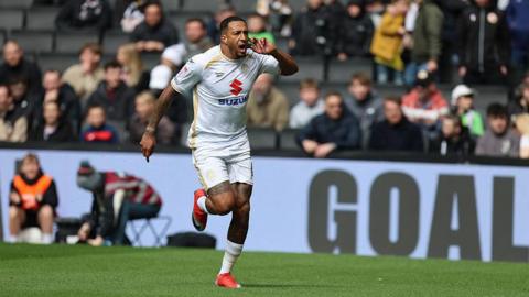Nathaniel Mendez-Laing of Milton Keynes Dons holds his left hand and finger to his ear as he runs on the pitch in celebration after scoring his side's first goal