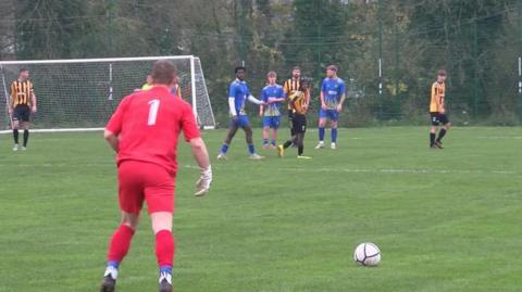 A scene from a football match with a goalkeeper in a red kit and with 1 on his back readying himself to take a free kick from inside his own half.