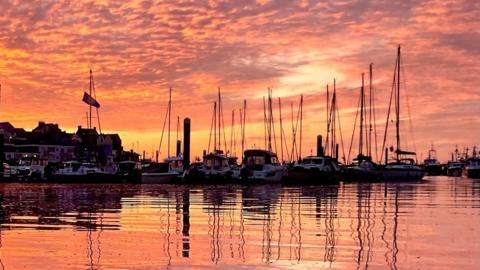 Boats and masts silhouetted against a red and orange sky which is reflected in the water