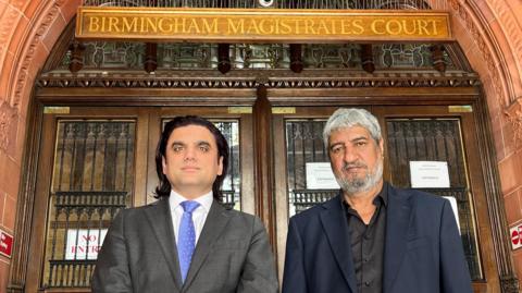 Iqbal Mohammed and Abdul Rehman stand on the steps of Birmingham Magistrates' Court standing side-by-side. They both wear suits. Behind them you can see the doors of the court and the red bricks in an ornate arch over the doorway. 