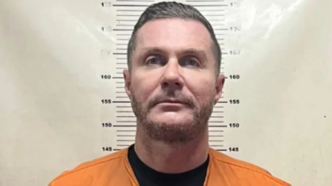Police mugshot of Steven Lyons. He is standing against a white height chart and staring directly at the camera. He has heavy stubble and short dark hair, combed in a side shed. He is wearing a black t-shirt and orange prison overalls.