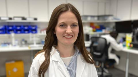 Sarah-Ann Ferguson is stood in the middle of the image smiling. She has long brown hair and is wearing a white lab coat and is standing in a lab.