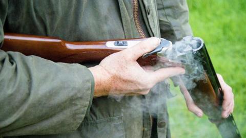The hands of a man wearing a green hunting coat grip a shotgun. The butt of the gun is tucked under his elbow and the open barrel hangs towards the grass. There are no cartridges in it and smoke rises from the empty chamber. 