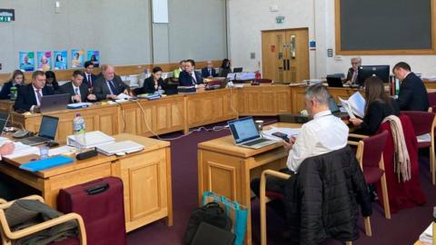 People wearing suits sit around big wooden desks in a council chamber