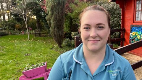 Healthcare assistant Emily Ballinger stands in a garden at Dove House Hospice, in front of a red shed, a pink wheelbarrow planter, green grass and shrubs. Emily has brown hair, tied back, and wears blue scrubs.