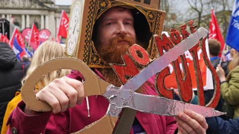 A man holds a cardboard placard that reads: NO MORE CUTS