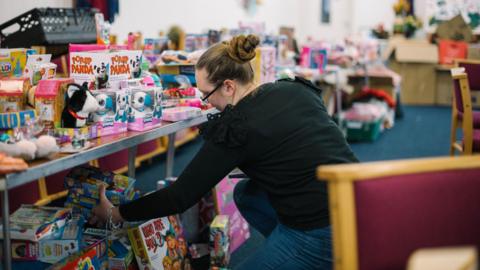 A woman wearing a black top and jeans holding several boxes of toys in one hand. The table in front of her is full of toys. 