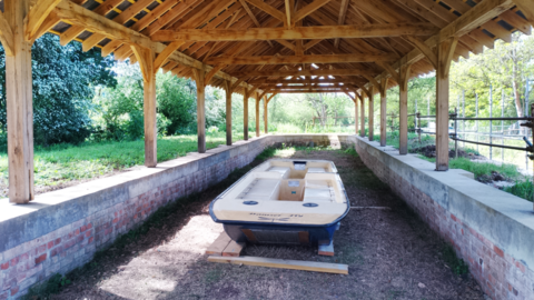 The hull of a narrowboat lying in the newly-restored dry dock,which has brick walls about two feet high with a wooden structure above with oak beams and a wooden roof. There is greenery all around