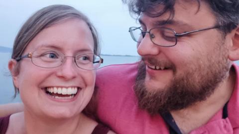 Alyssa and her husband are hugging, with a coastal view behind them. They are both smiling and she is looking off to the right, while he is looking at his wife.