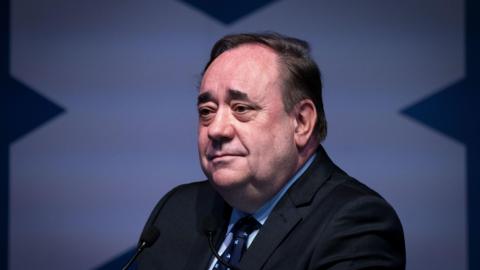Former Scottish first minister Alex Salmond is wearing a dark suit and blue tie patterned with white saltire crosses, against a blue and white background.