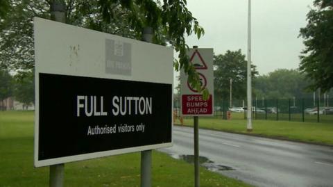Sign at the entrance to HMP Full Sutton displaying the words “FULL SUTTON” and “Authorised visitors only”. The sign stands next to a road bordered by grass and trees. A fence and prison grounds are visible in the background under an overcast sky.