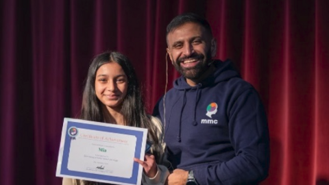 Mia is standing next Atwal holding a certificate and smiling. She has long, black hair and has a brown cardigan on over a black top. Atwal is wearing a blue hoodie and is smiling at the camera. They stand in front of a maroon curtain.