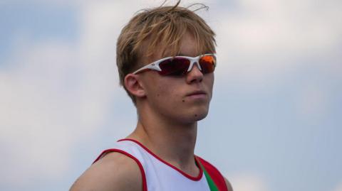 A young man with blond hair wearing white sunglasses and a white Jersey. Behind him is blue sky with white clouds.