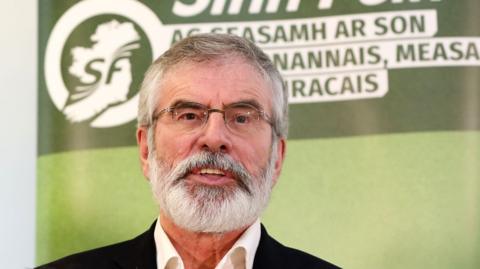 Gerry Adams, standing infront of a green Sinn Fein poster, pictured in 2017. He has a short grey beard and grey hair.