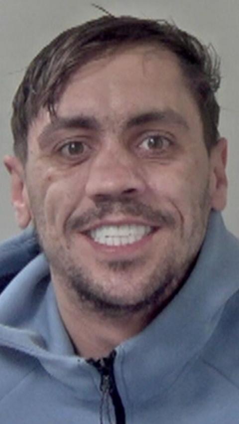 A man in a blue tracksuit with white teeth in a police mugshot.