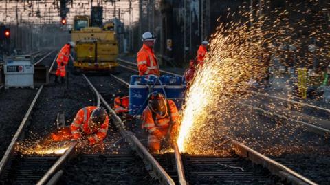 Workers in orange clothing and white hard hats work on a railway line at dusk. They are wearing safety helmets as a jet of bright sparks flies from the track they are using tools on.