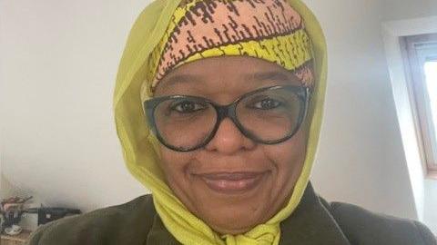 Zainab Nasko, who is wearing a yellow headscarf, black glasses and a green blazer, smiles for a photograph in an indoor area by a window.