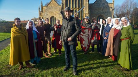 A group of people stand on grass in front of an old stone church, dressed in medieval-style clothing and armour. In the centre, a man in modern outdoor clothes and a hat smiles at the camera, while others around him wear long robes, cloaks, helmets, and carry swords and a red shield with a white lion emblem.