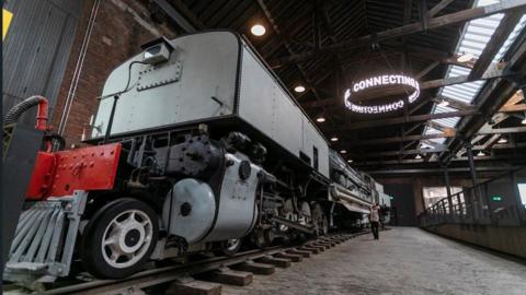 Inside the Power Hall is a large grey locomotive. A woman is staring up at the vehicle which is housed within the red-bricked warehouse. 