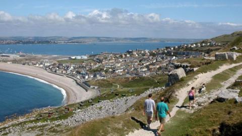 View across Portland Harbour and Chesil Beach from the top of the hill on Portland on a sunny day. In the foreground is a group of three walkers and a dog. At the bottom of the hill is the town of Fortuneswell, sandwiched between Chesil Beach and Portland Harbour. Weymouth and the Dorset coast can be seen across the bay in the distance.