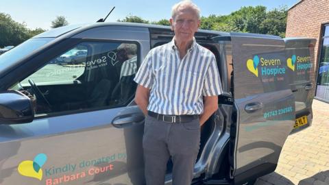 An older man with white hair is standing in front of a grey van with blue and yellow writing on it. On the passenger door, writing reads "Kindly donated by Barbara Court". The man is wearing a blue and white striped top with grey trousers