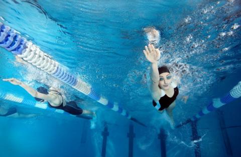 Two women in black swimming costumes and caps swimming in a pool, seen from below