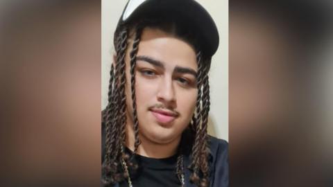 Mason Miller had long dark brown hair in twisted plaits, and a moustache. He is wearing a black and white baseball hat, a black T-shirt and chain.