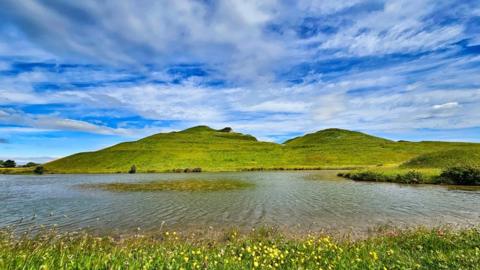 Blue sky with streaks of cloud sit above two green mountains with a lake in front and grasses in the foreground