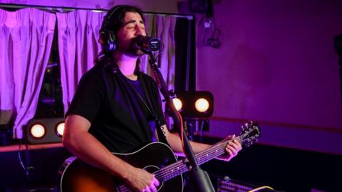 Noah Kahan in the BBC Radio 1 Live Lounge. Noah, 26, has brown hair down to his shoulders and a short brown beard. He wears a black T-shirt and is playing an acoustic guitar. The live lounge is lit by a purple light and behind him you can see sound equipment, lights and curtains. He stands in front of a microphone and sings into it, his eyes closed.