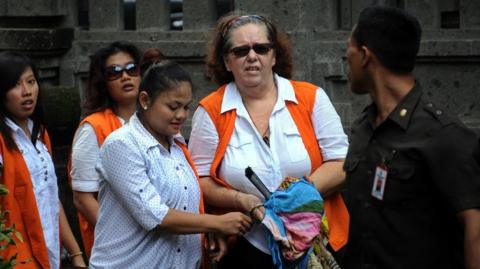 Lindsay Sandiford, who has short curly hair, walking alongside Indonesian officials. One is dressed in a green uniform and the others are wearing white blouses with orange tops.