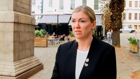 Deputy Raluca Kovacs, a woman with blonde hair which is tied back. She is wearing a black blazer and white top. She has two badges on her lapel. She is standing in Jersey's Royal Square, in front of the entrance to the Cock and Bottle pub. There are orange, autumnal leaves on the ground beneath the trees in the square. 