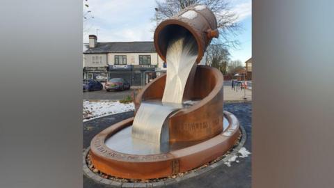 A sculpture stands in front of a row of shops. The sculpture depicts a brown vessel suspended in the air and pouring out a silver river of metal. The silver strip flows into another brown vessel on the ground. There is a small amount of snow on the street in the background.