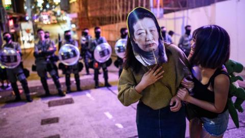 A woman wears a face mask depicting Secretary for Justice Teresa Cheng as she poses for photos in front of a line of riot police as they stand in a line to block entry to the Lan Kwai Fong area during Halloween in Hong Kong on October 31, 2019