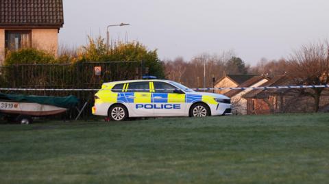 A police car behind a police cordon on a residential street by a patch of grass