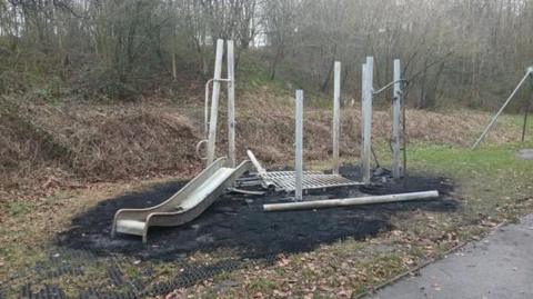 The remains of the damaged play equipment. Most of the metal slide is lying on the ground. Seven metal supports remain upright but the equipment is unusable. The rubber protective mat surrounding the slide has melted. Trees and shrubs stand behind the equipment in the park.